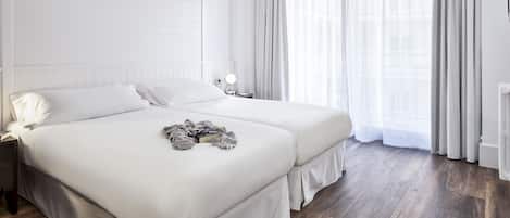 A modern hotel room with two beds, a large window with sheer curtains, a bedside table with a lamp, and a radiator.