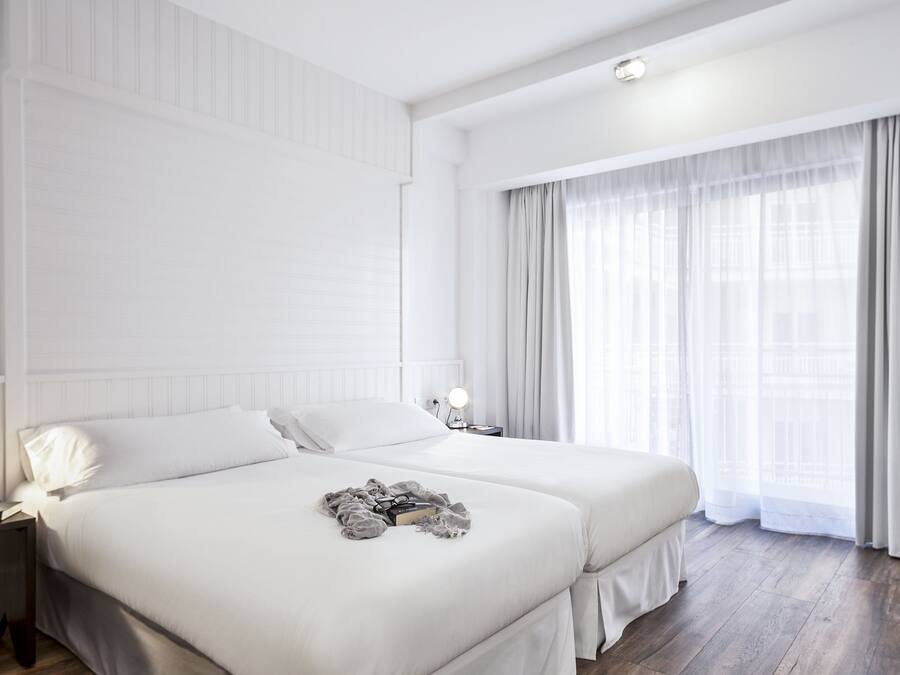 Habitación de hotel moderna con dos camas, un ventanal grande con cortinas translúcidas, una mesita de noche con lámpara y un radiador.