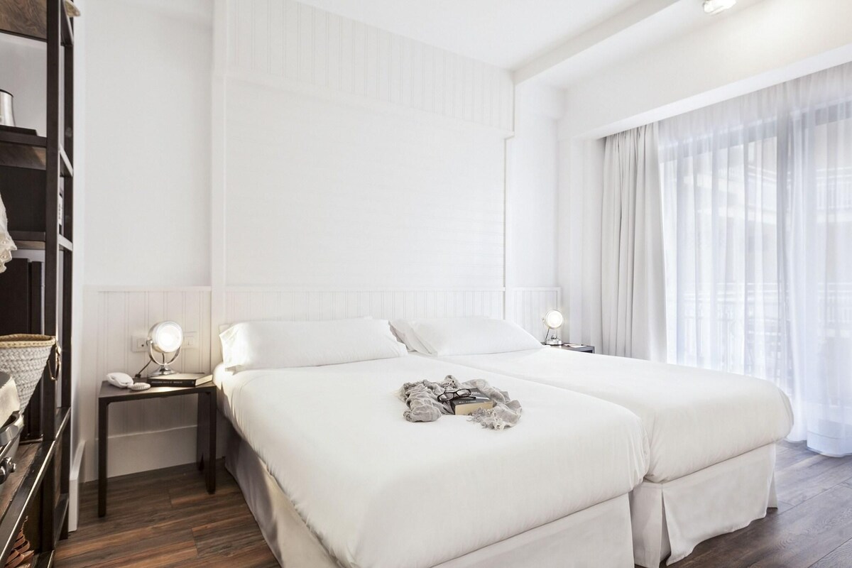 Une chambre d’hôtel avec un grand lit, une table de chevet avec une lampe et une fenêtre avec des rideaux légers.