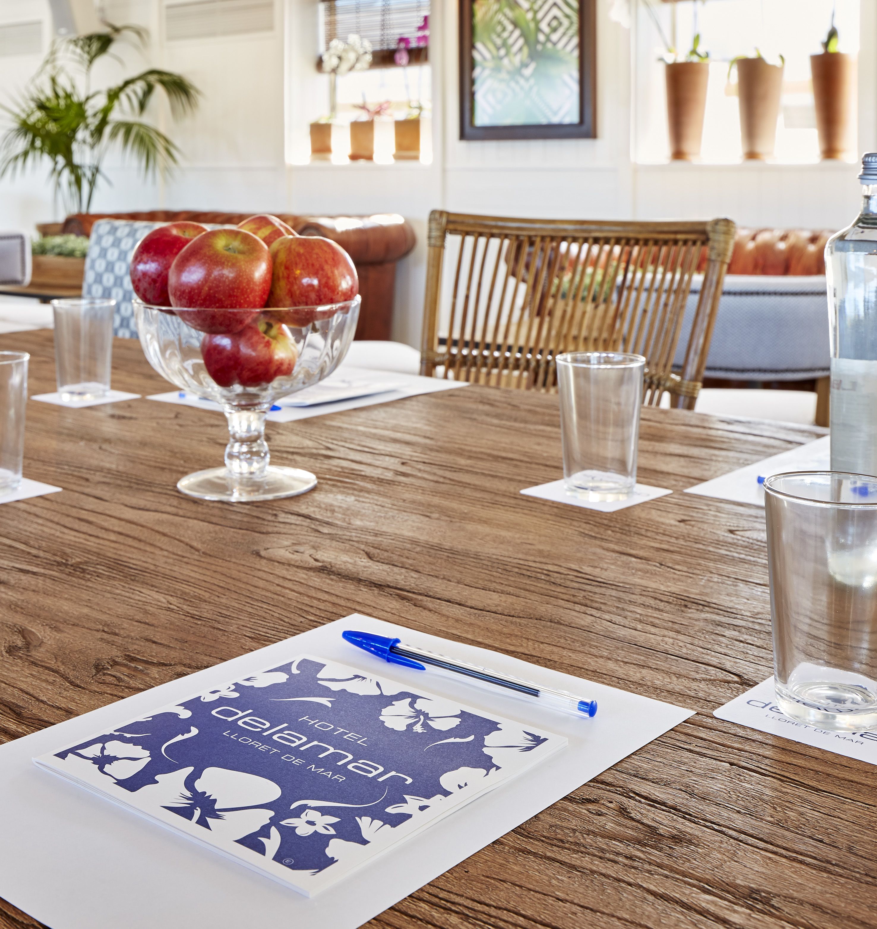 Una mesa de madera con un cuenco de vidrio lleno de manzanas, un bolígrafo y un folleto de hotel.