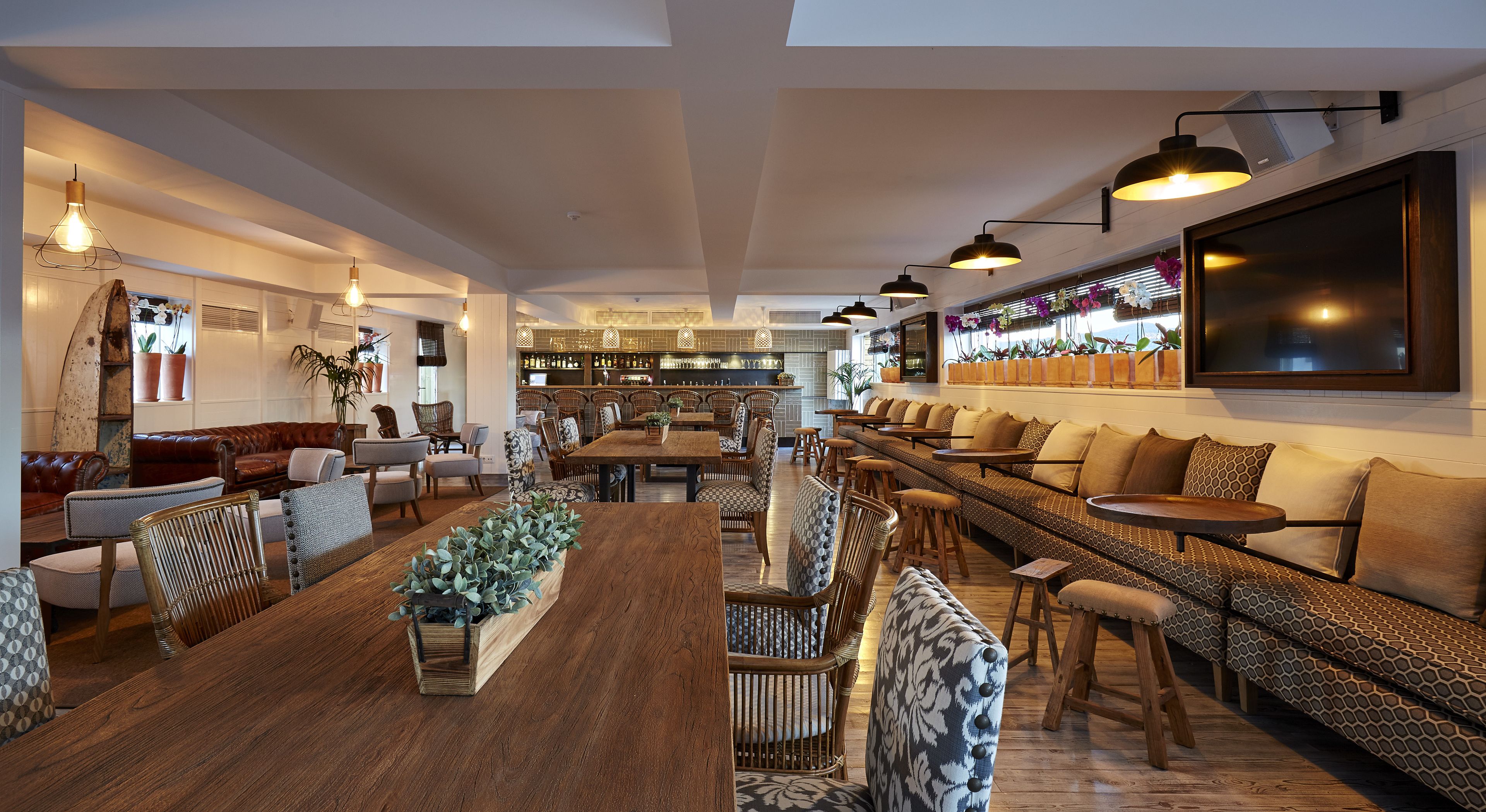 A spacious, well-lit lounge area with a long wooden table, comfortable seating, and a bar area in the background.