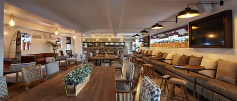 A spacious, well-lit lounge area with a long wooden table, comfortable seating, and a bar area in the background.