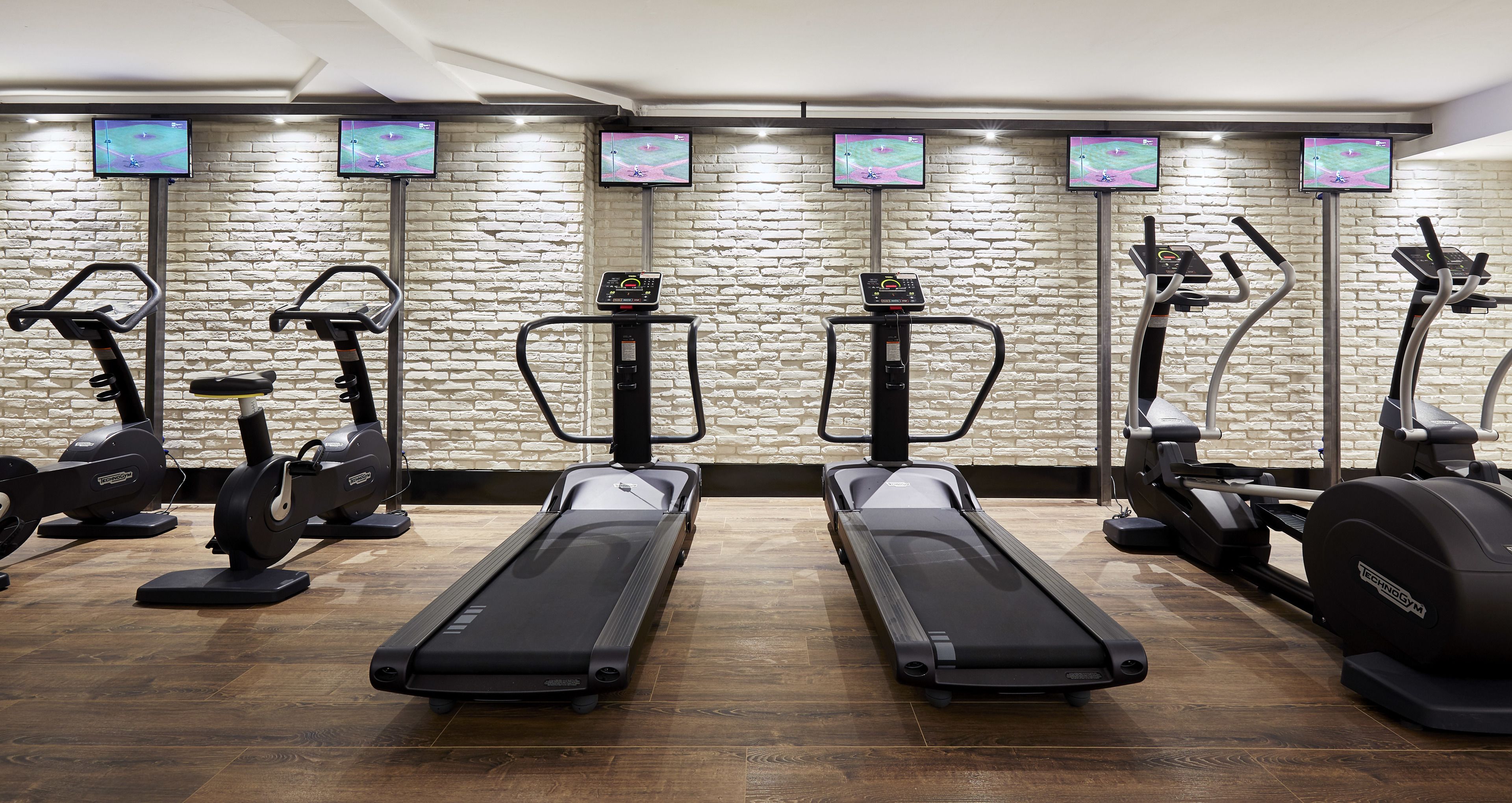 Un gimnasio con cintas de correr, elípticas y bicicletas estáticas, equipado con pantallas digitales y un fondo de pared de ladrillo.