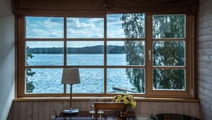 A room with a large window offering a view of a lake and forest. There is a table with a lamp, a chair, and a vase with flowers. - Porto Resort (Lilaste)