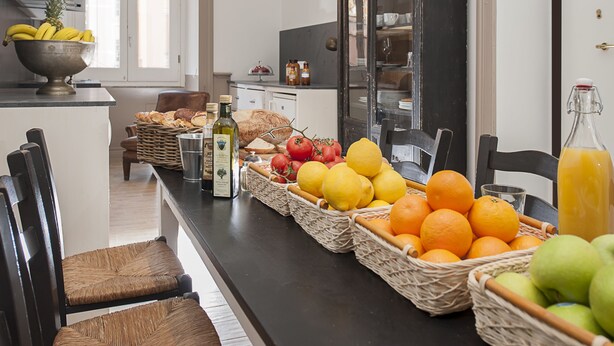 A kitchen with a long countertop filled with fruits and vegetables, a bread basket, and a bottle of olive oil. - Côté Rome Colosseo (Rome)