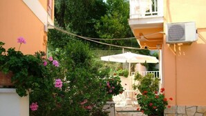 Un patio estrecho empedrado con plantas y flores en macetas, un paraguas blanco y una silla blanca.