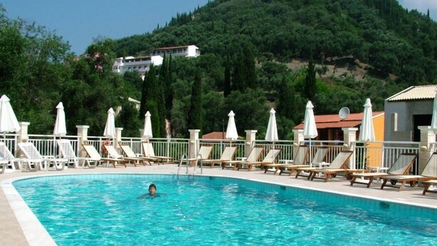 Una piscina con sillas de descanso y sombrillas, rodeada de vegetación y edificios al fondo.