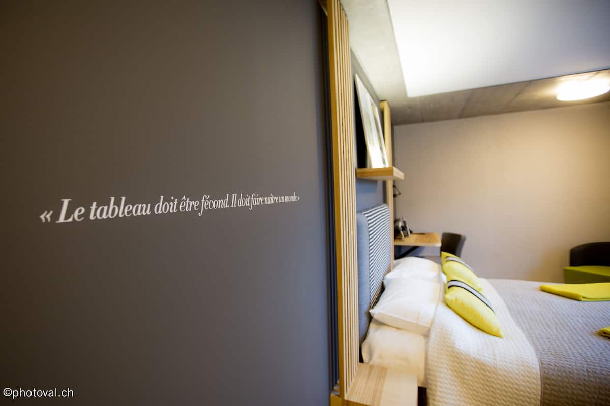 A hotel room with a bed, a bedside table, a wall with a quote, and a ceiling light.