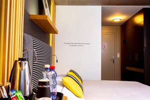Habitación de hotel con cama, cafetera, una botella, almohada y una pared con una cita.
