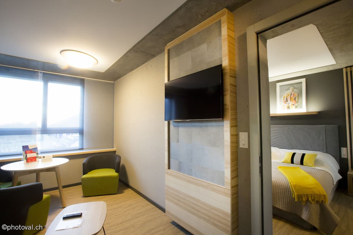 A modern hotel room with a bed, a small table, and a chair. There is a window providing a view outside.