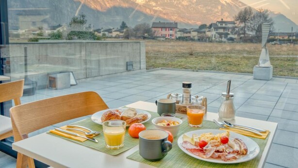 A breakfast table with various food items and drinks, set against a backdrop of mountains.