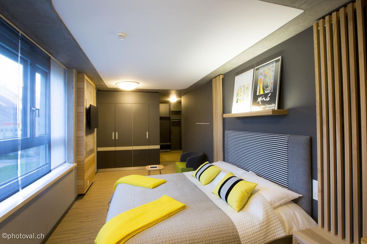 A modern hotel room with a large bed, a small seating area, and a view of the outdoors.