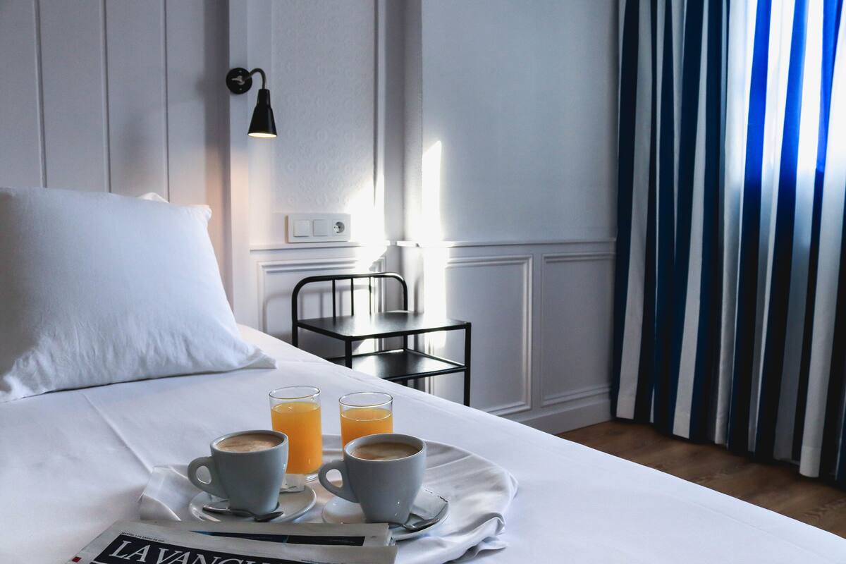 Habitación de hotel con una cama, una bandeja con dos tazas de café y dos vasos de zumo de naranja, un periódico, una mesita con una silla y una ventana con cortinas azules.
