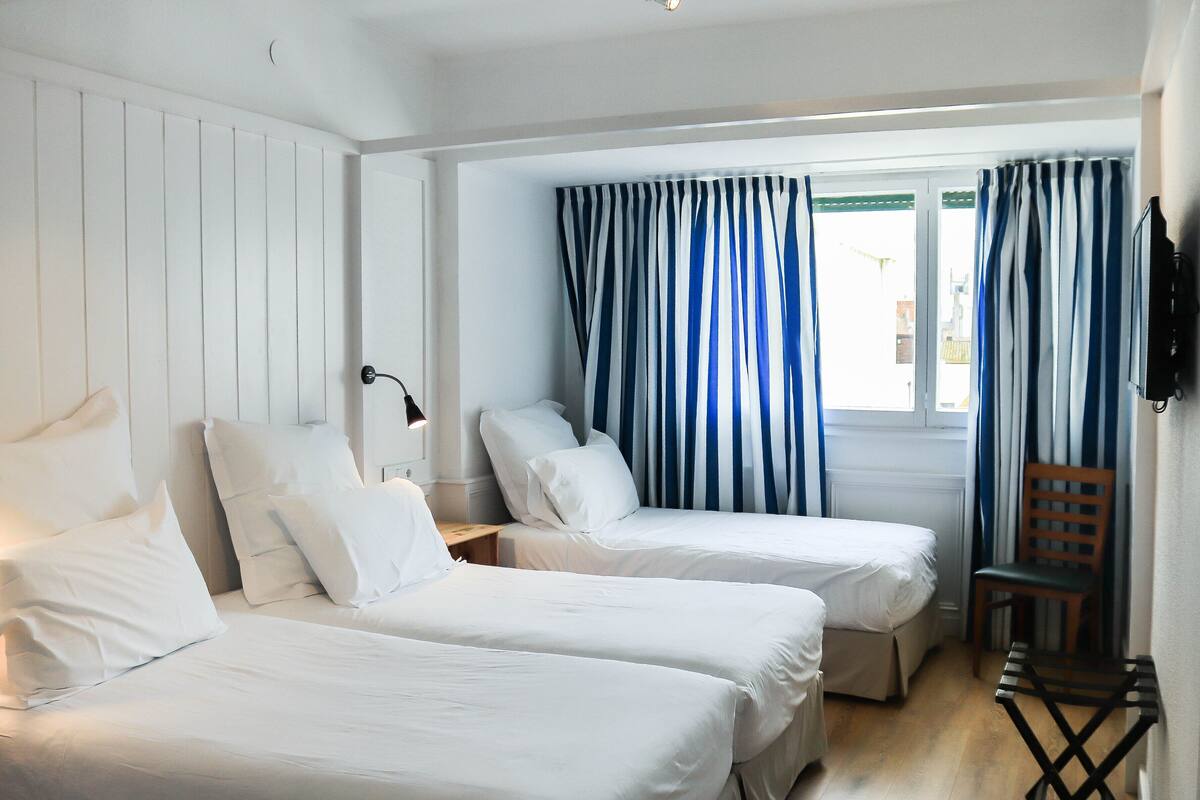 Una habitación de hotel con dos camas individuales, una silla de madera, un escritorio con lámpara y una ventana con cortinas a rayas.