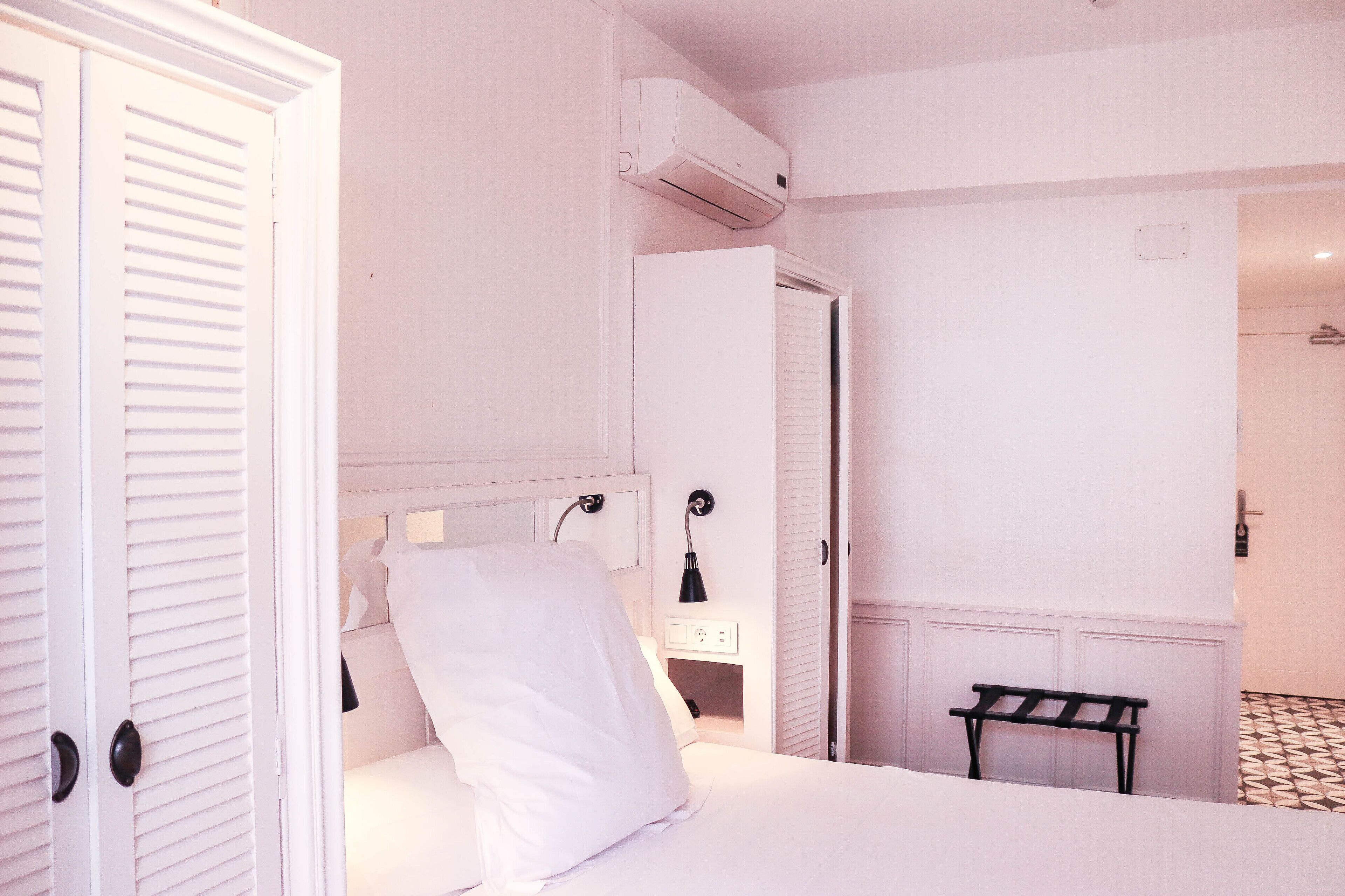 A bedroom with a bed, a bedside table, a lamp, a bench, and an air conditioning unit.