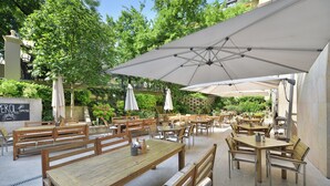 An outdoor café with wooden tables and benches, umbrellas, and greenery in the background. - Wilson Palace by LOFT (Bratislava)
