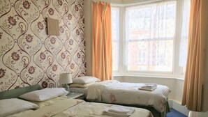 Quarto de hotel com duas camas, uma mesinha de cabeceira, uma luminária e uma janela com cortinas.