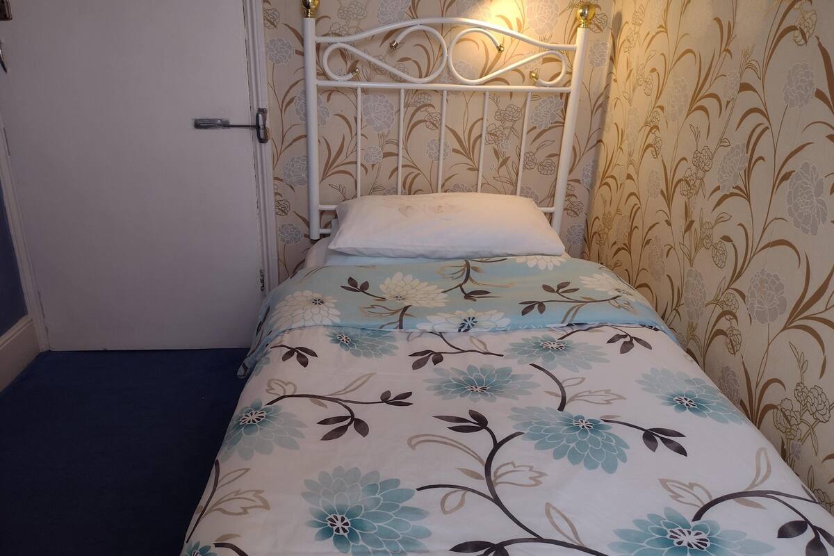 A small, single-bed room with floral wallpaper, a white metal headboard, and a bedside lamp.