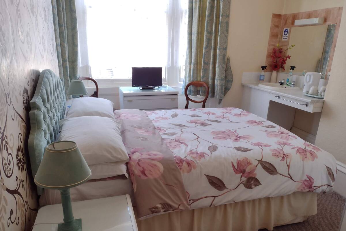 A bedroom with a bed, bedside table, lamp, chair, desk with a computer, and a bathroom with a sink and mirror.