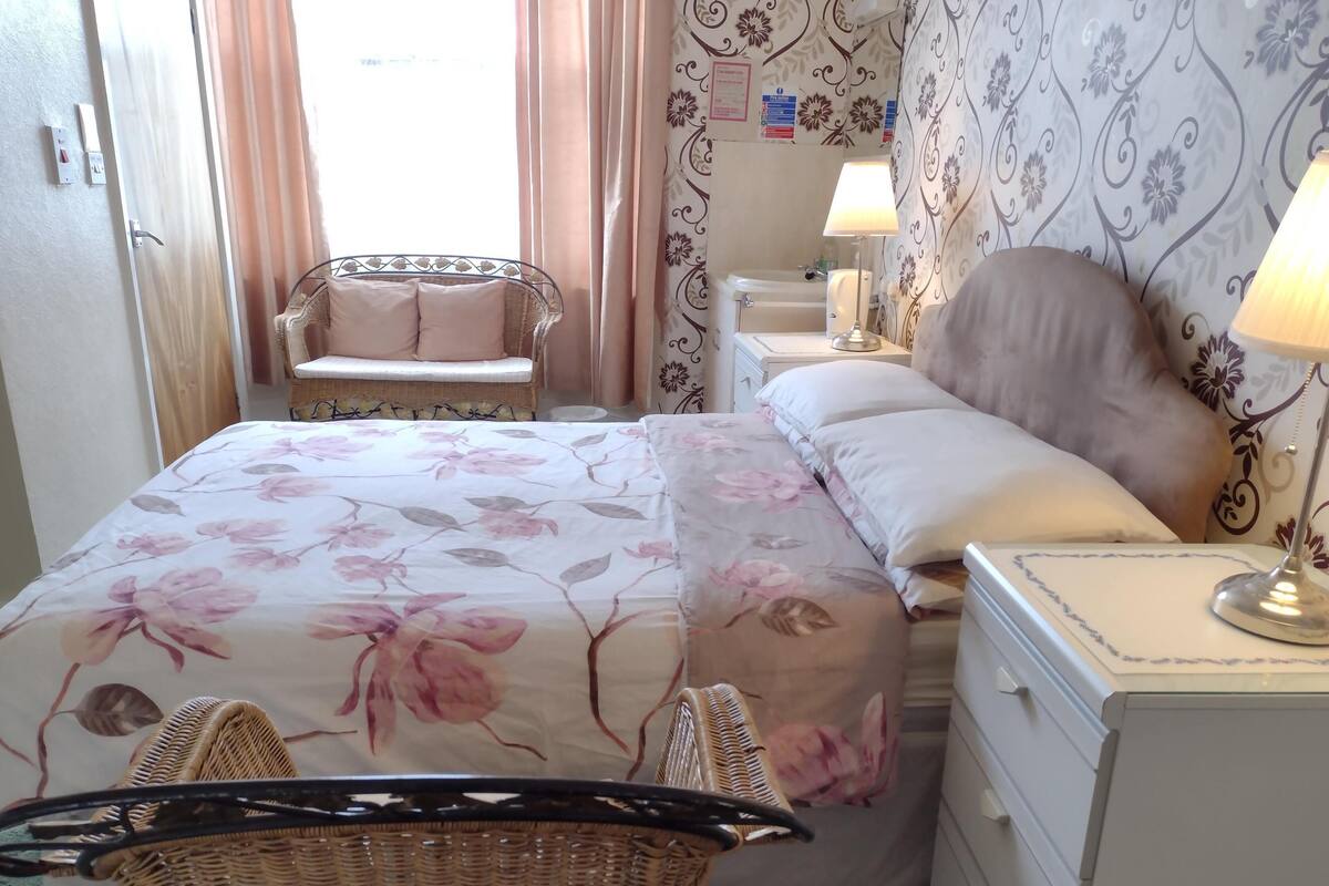 A bedroom with a bed, a nightstand, a lamp, a wicker chair, and a window with curtains.