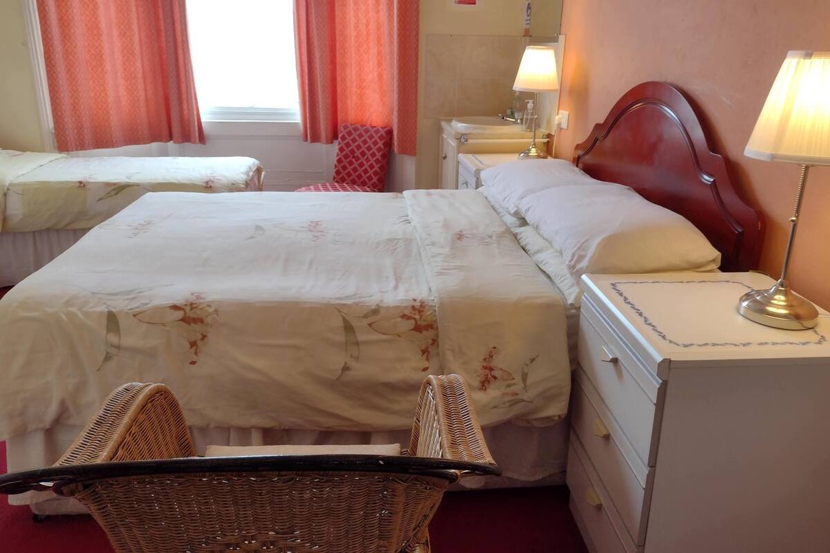 A hotel room with two beds, a wooden headboard, a bedside lamp, a wicker chair, and a small table.