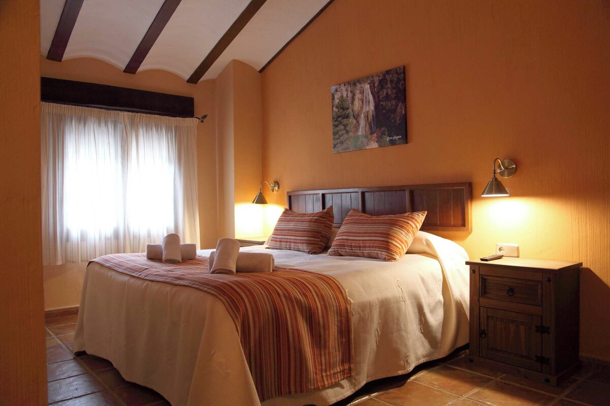 A bedroom with a large bed, a nightstand, a wall-mounted lamp, and a painting.