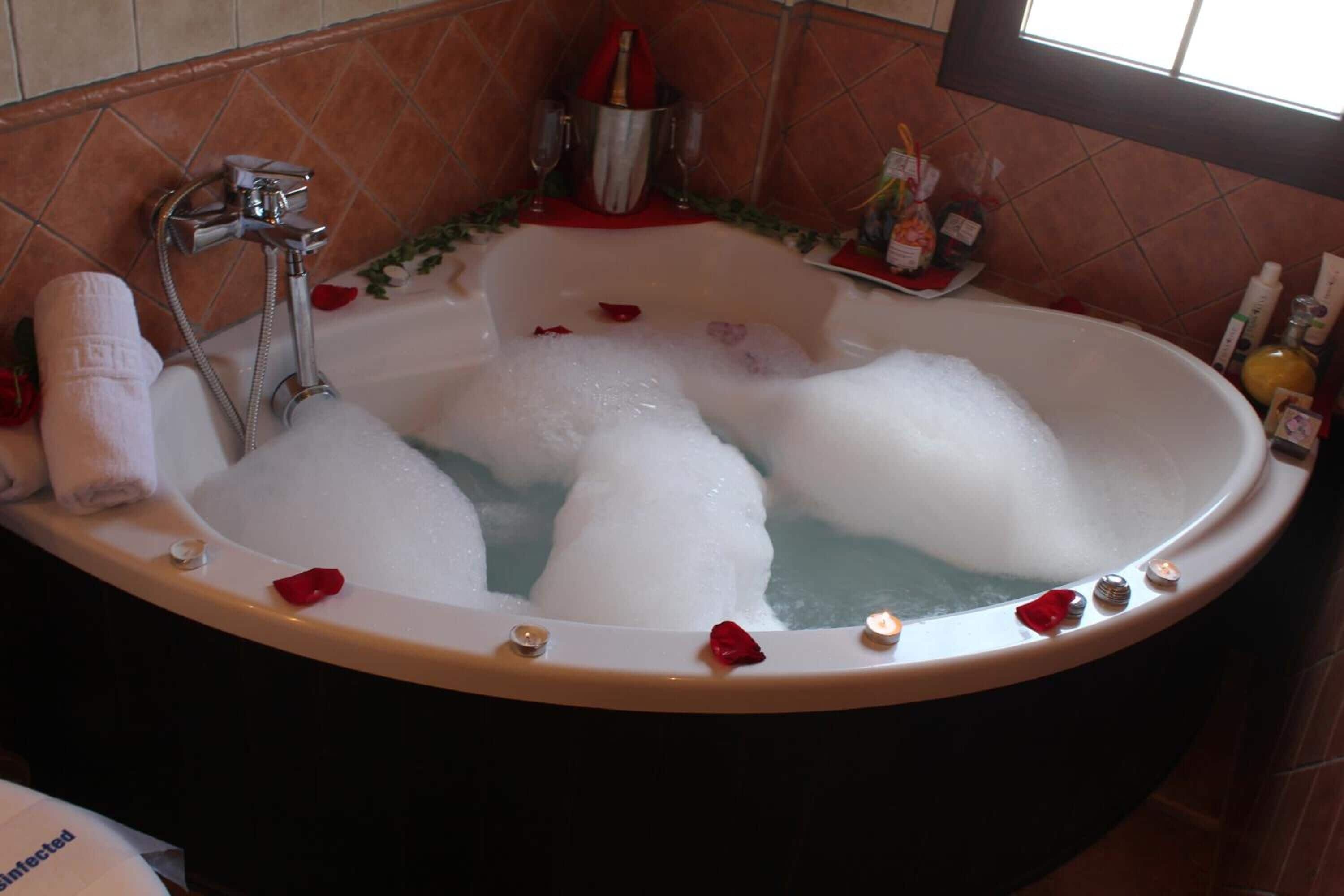 Una bañera llena de burbujas, rodeada de pétalos de rosa y velas, con una toalla y una botella de champán.