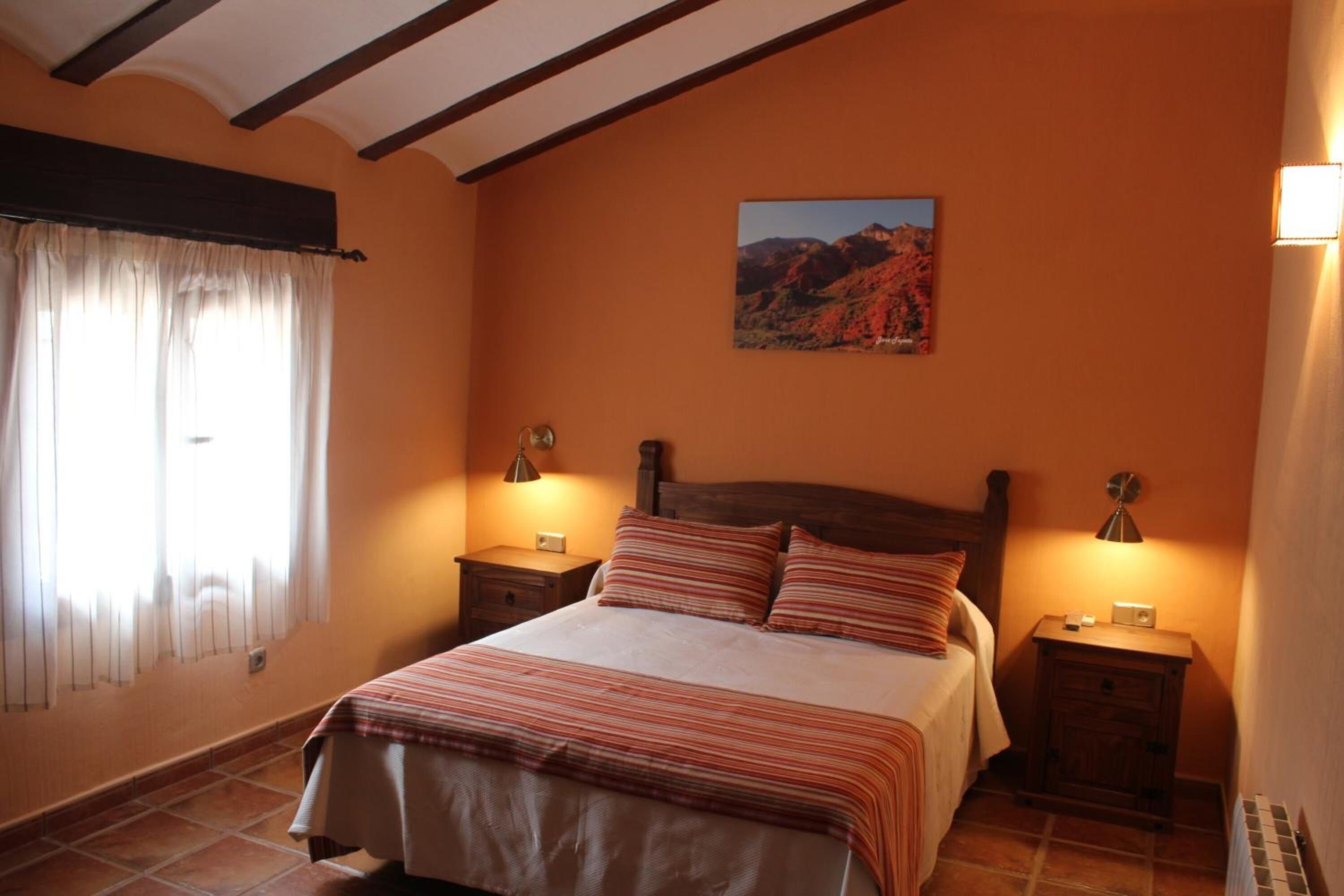 A bedroom with a bed, two bedside tables, a nightstand, a window with curtains, and a wall-mounted picture.