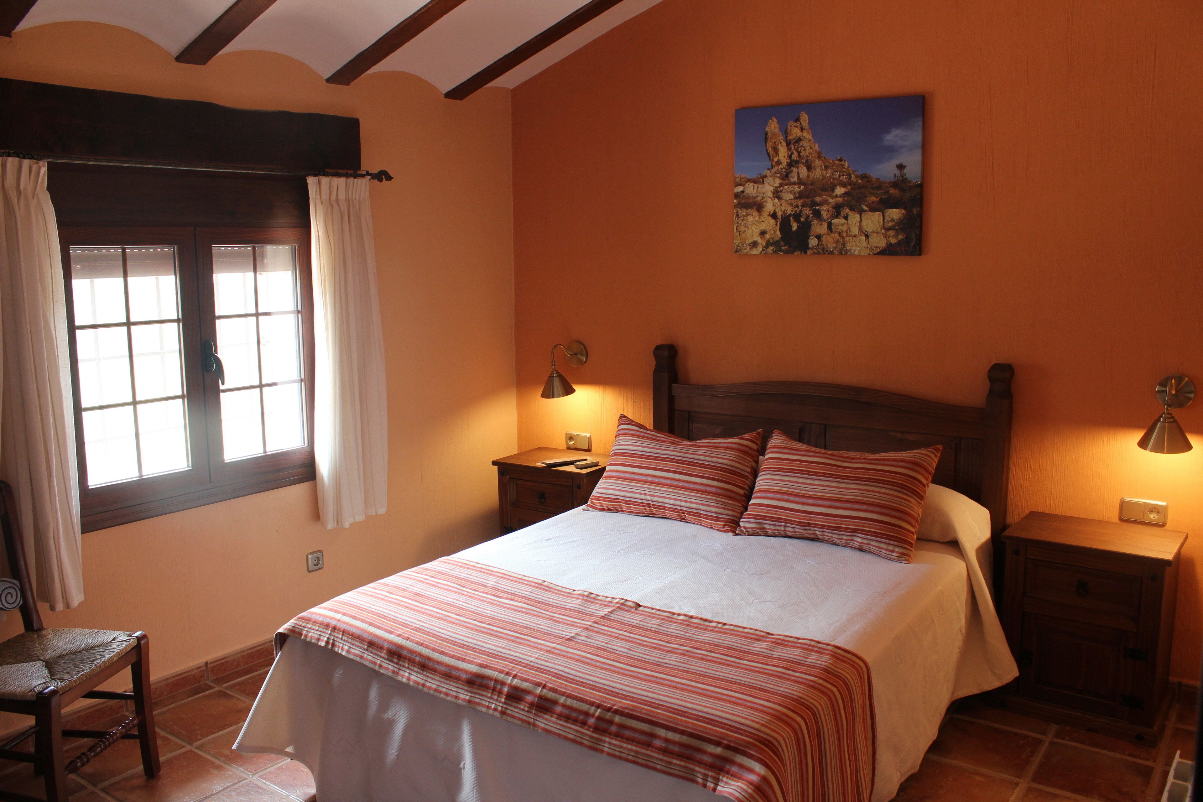 Un dormitorio con cama, dos mesitas de noche, una lámpara, una silla, una ventana con cortinas y un cuadro en la pared.