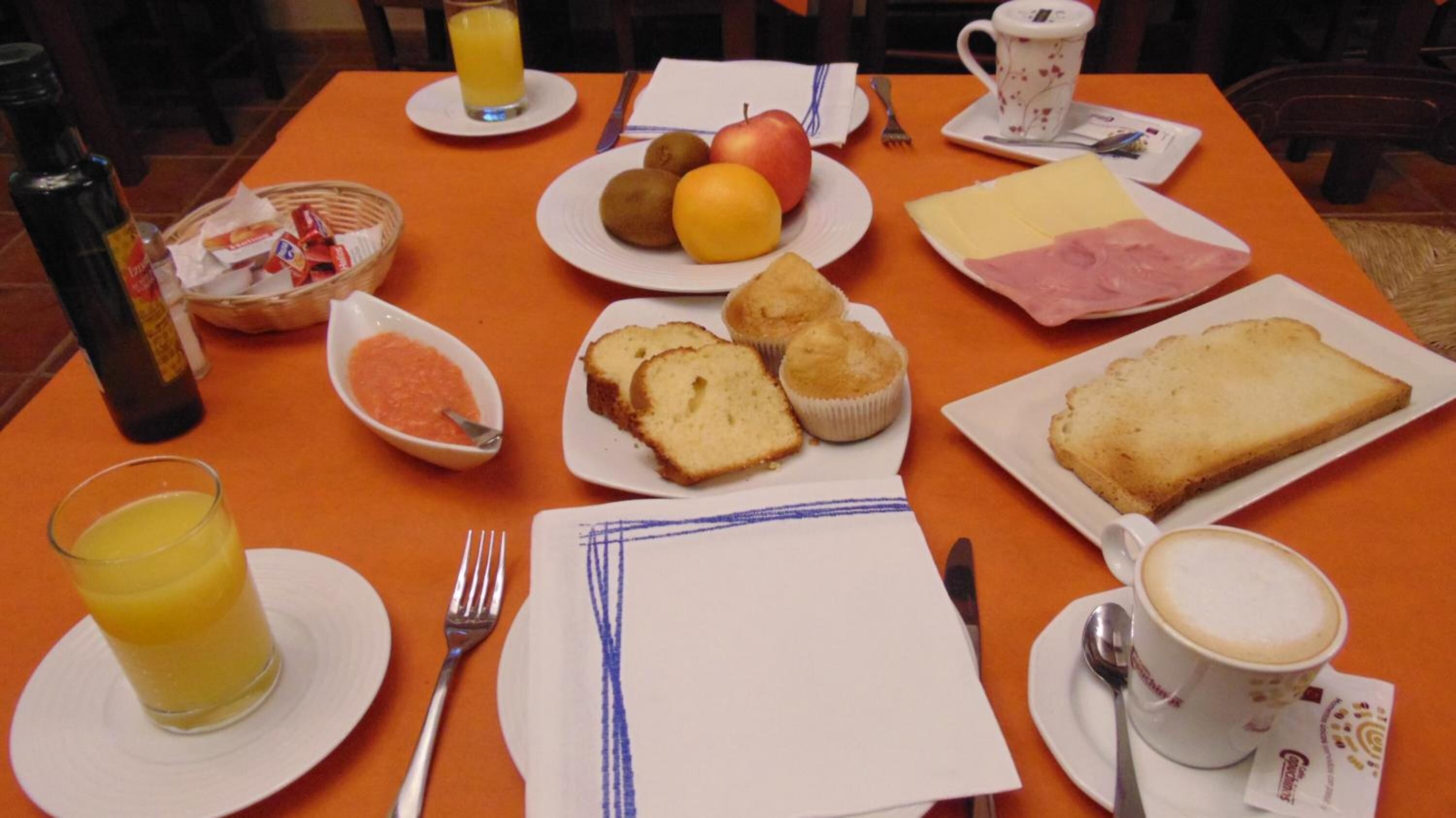 A breakfast table with plates of toast, muffins, cheese, ham, fruits, and a cup of coffee.