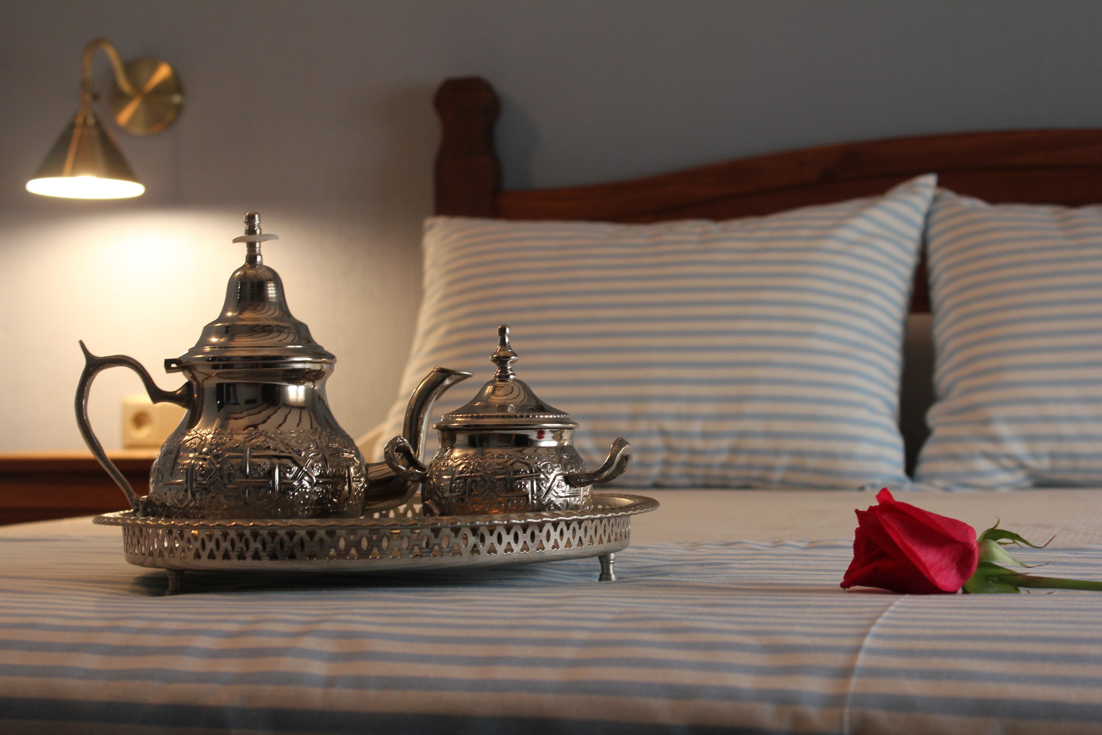 Ein Bett mit einem Metalltablett, das zwei kunstvolle Teekannen, eine einzelne Rose und eine Wandlampe beherbergt.