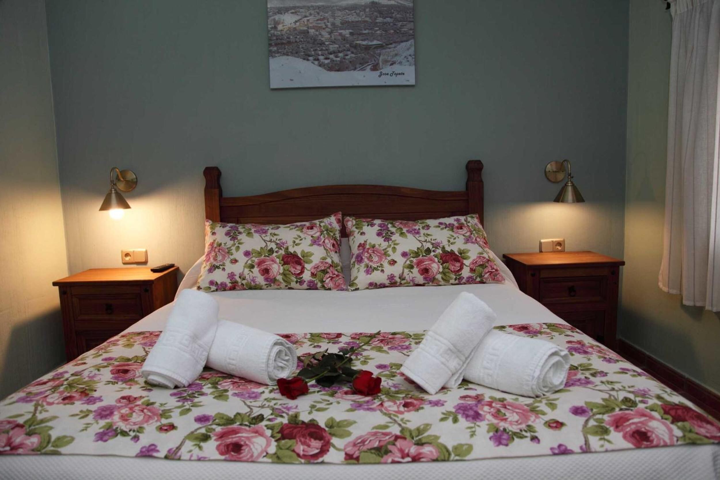 Un letto rifatto con cura, con lenzuola a motivi floreali, due lampade sui comodini e un quadro con una veduta cittadina appeso al muro.