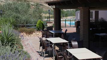 Un patio al aire libre con mesas y sillas, una piscina al fondo y un paisaje montañoso.