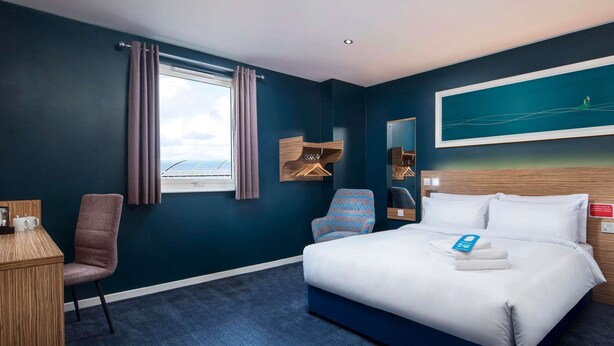 A hotel room with a bed, a desk, a chair, a window with curtains, a painting on the wall, and a view of the sky. - Travelodge Ryde Isle of Wight (Ryde)