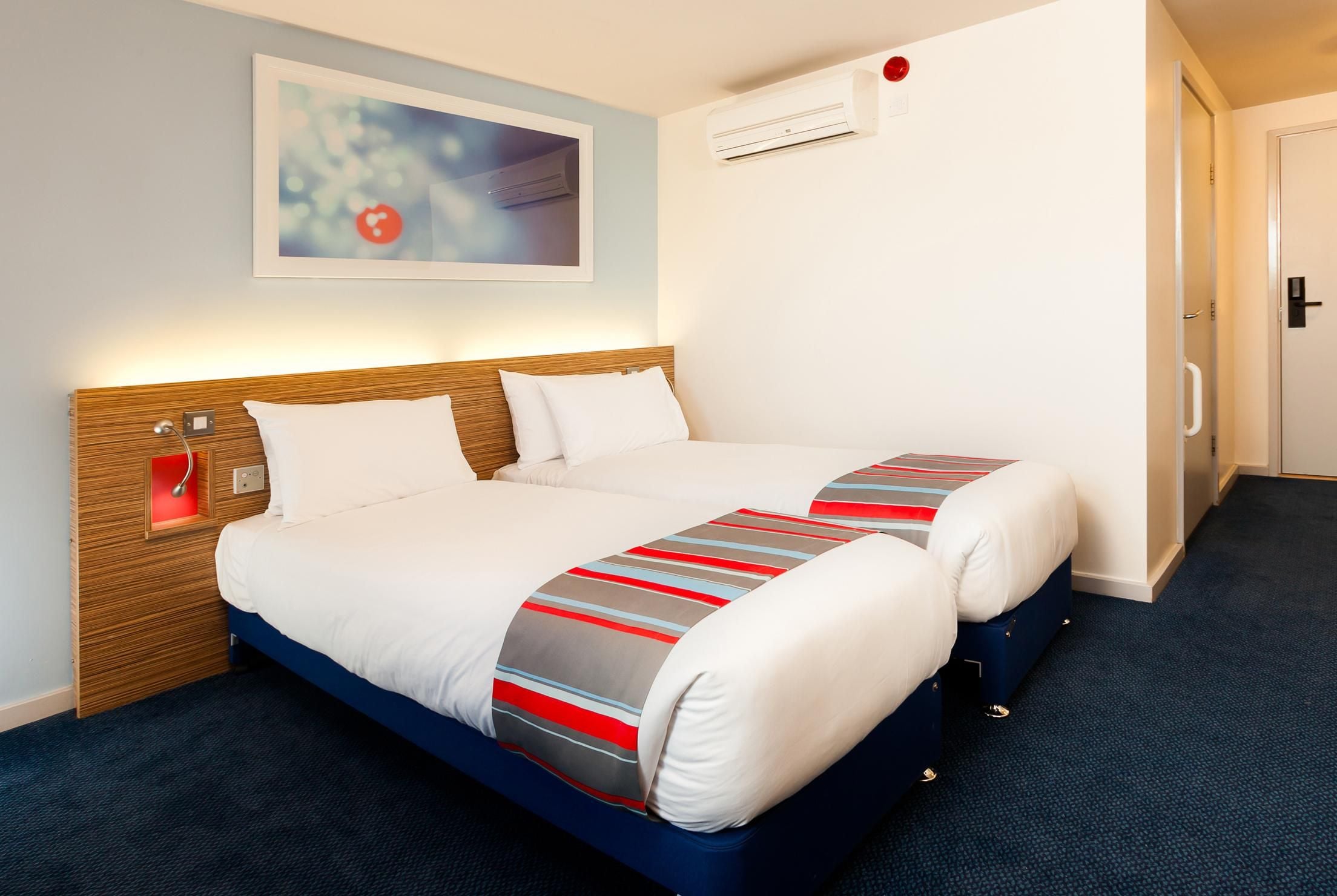 A hotel room with a bed, bedside controls, an air conditioning unit, and a door.