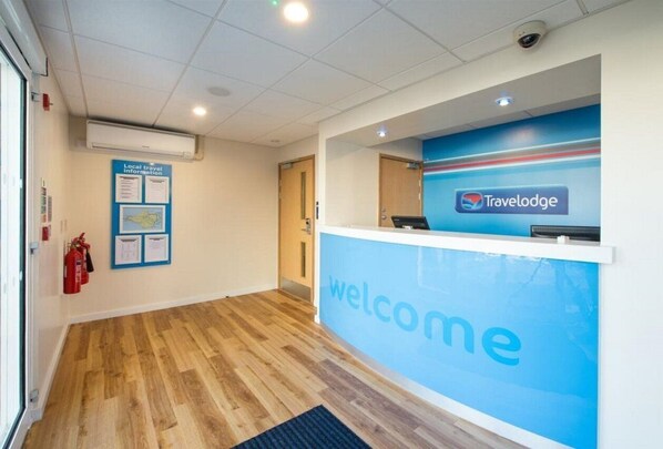 Uma área de recepção com uma placa azul que diz 'Welcome', o logotipo 'Travelodge' e um quadro de informações turísticas locais.