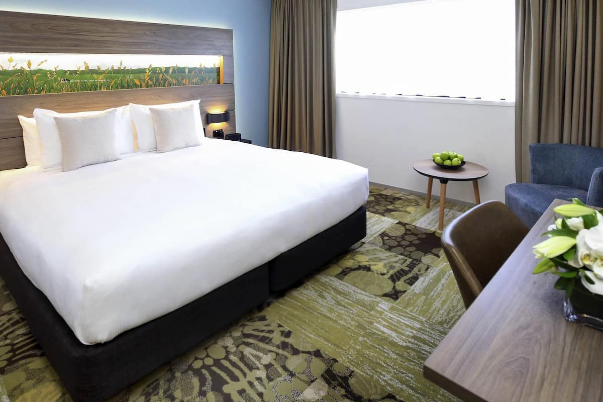 A hotel room with a large bed, a small table with fruit, a chair, and a view of a field through a window.