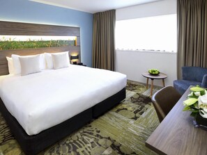 Habitación de hotel con una cama grande, una mesita con fruta, una silla y vistas a un campo por la ventana.