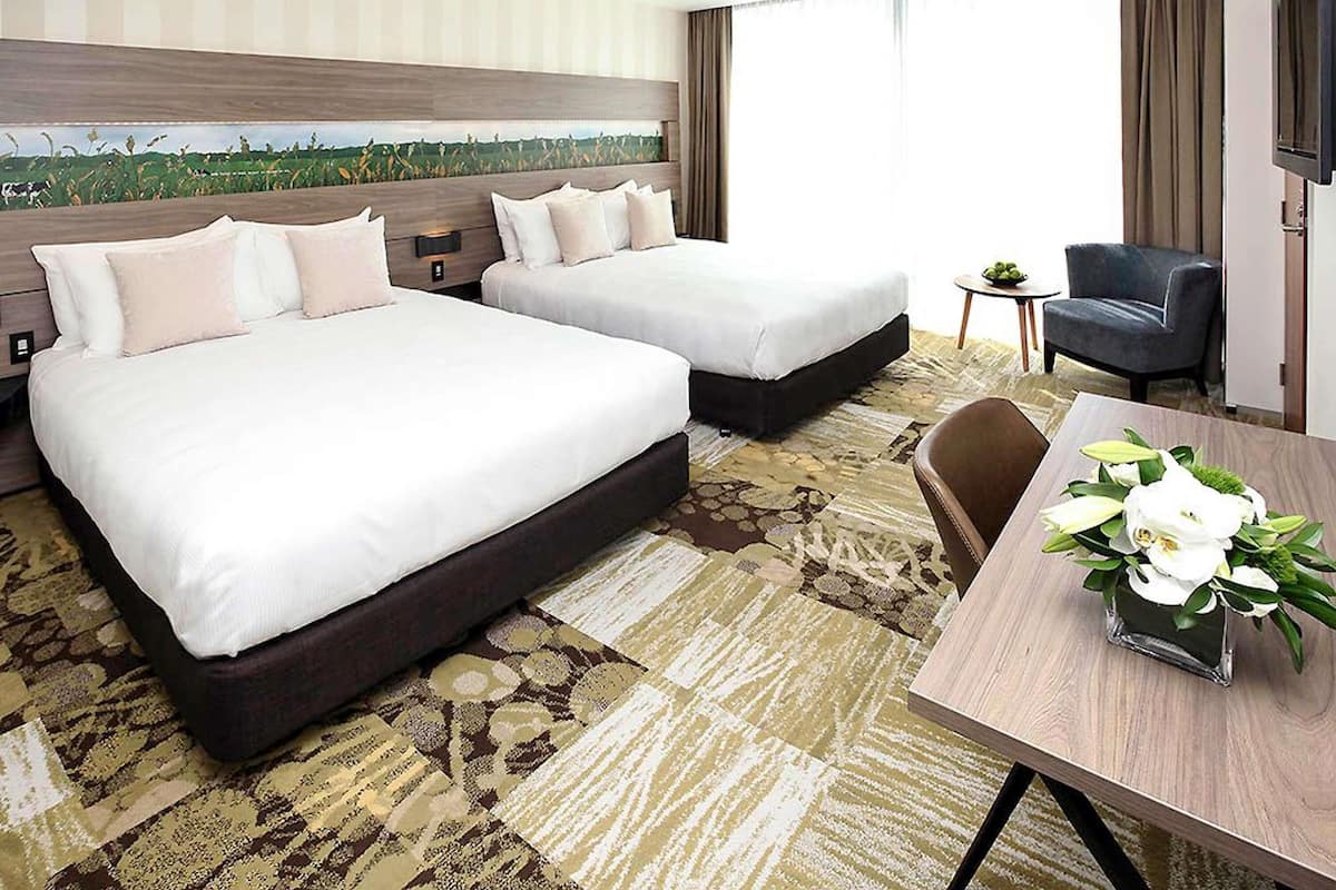 A hotel room with two beds, a TV, a small table with a vase of flowers, and a view of a field through the window.