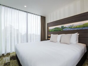 A hotel room with a large bed, a headboard with a landscape painting, and sheer curtains.