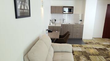 A modern living room with a beige sofa, a small round wooden table, and a patterned carpet. A kitchen with a microwave and cabinets is visible in the background.