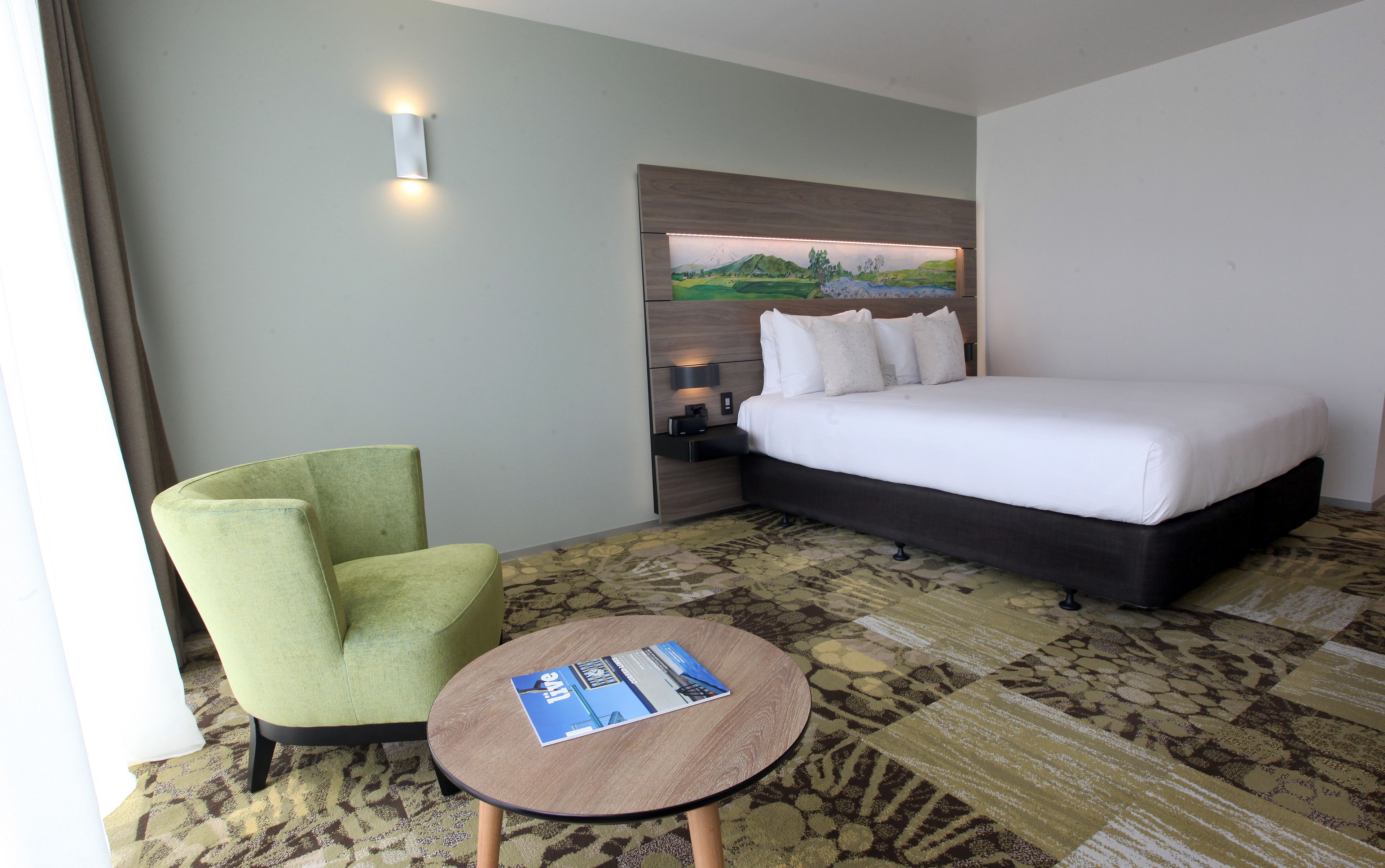 Une chambre d’hôtel avec un lit, un fauteuil vert, une petite table et un tableau au mur.