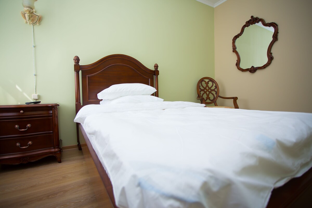 A neatly made bed with white linens, a wooden nightstand with drawers, and a chair with a decorative backrest.