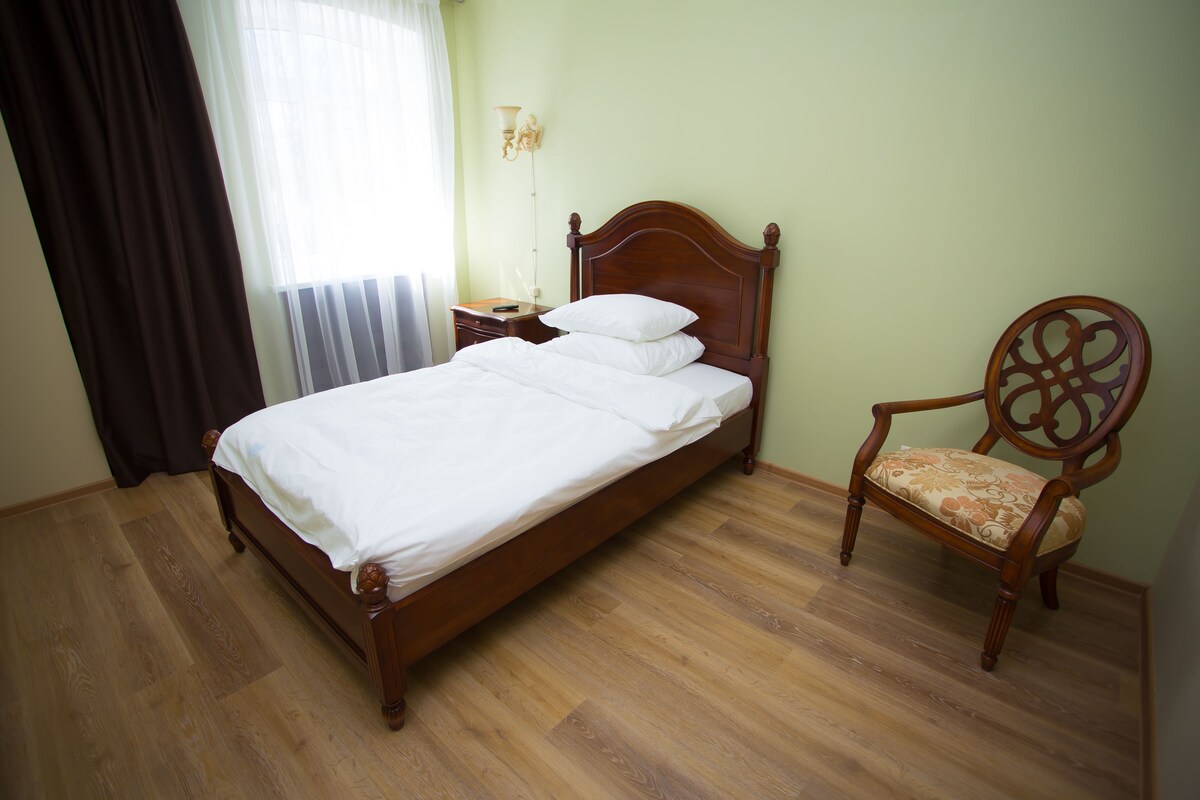 A neatly made bed with white linens, a wooden nightstand with a lamp, and a wooden chair with a floral cushion.