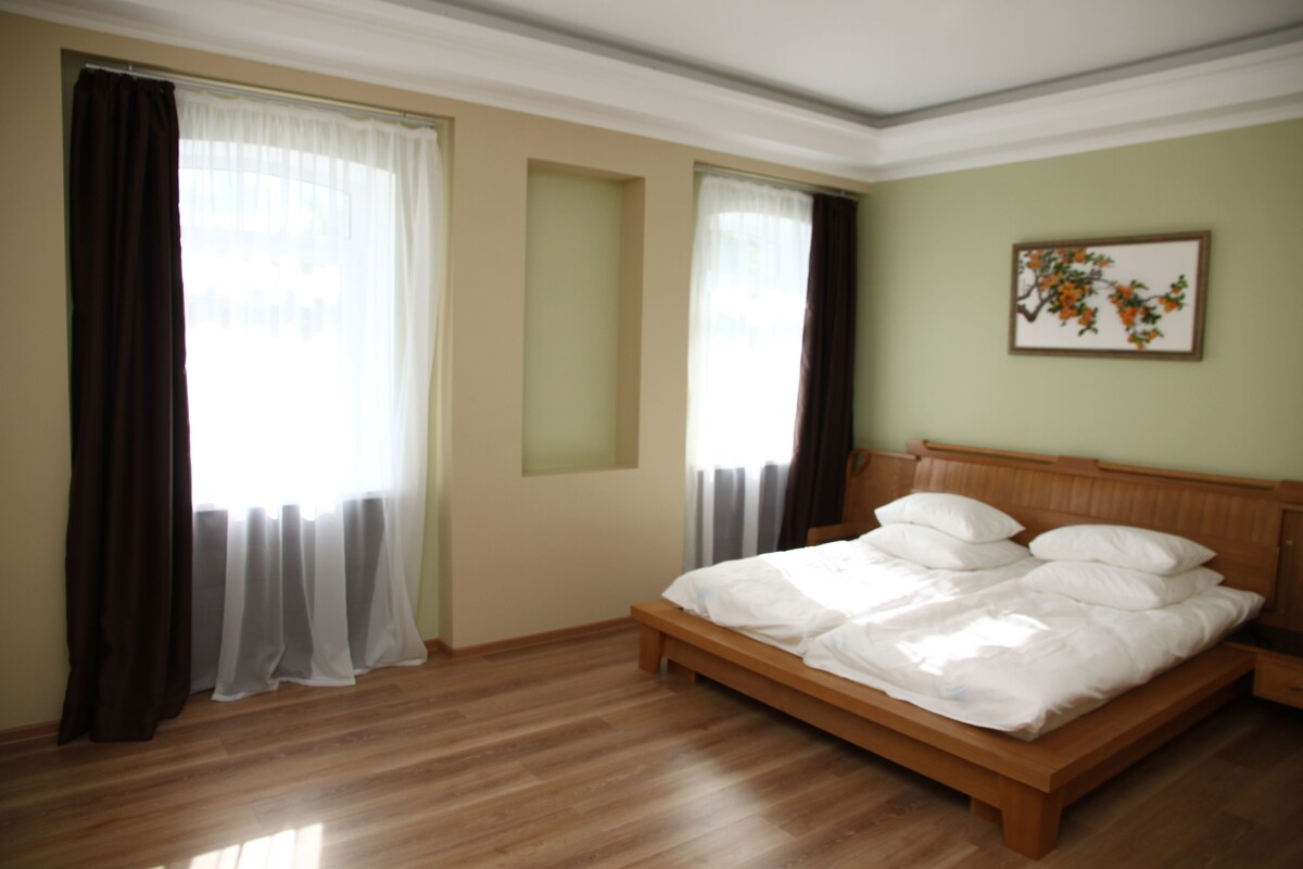 A bedroom with a wooden bed, two windows with sheer curtains, a nightstand, and a wall-mounted picture.