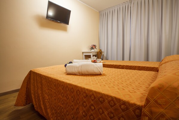 A hotel room with two beds, a flat-screen TV mounted on the wall, a small table with a vase of flowers, and a window with curtains. - Hotel Olimpic (Sestriere)