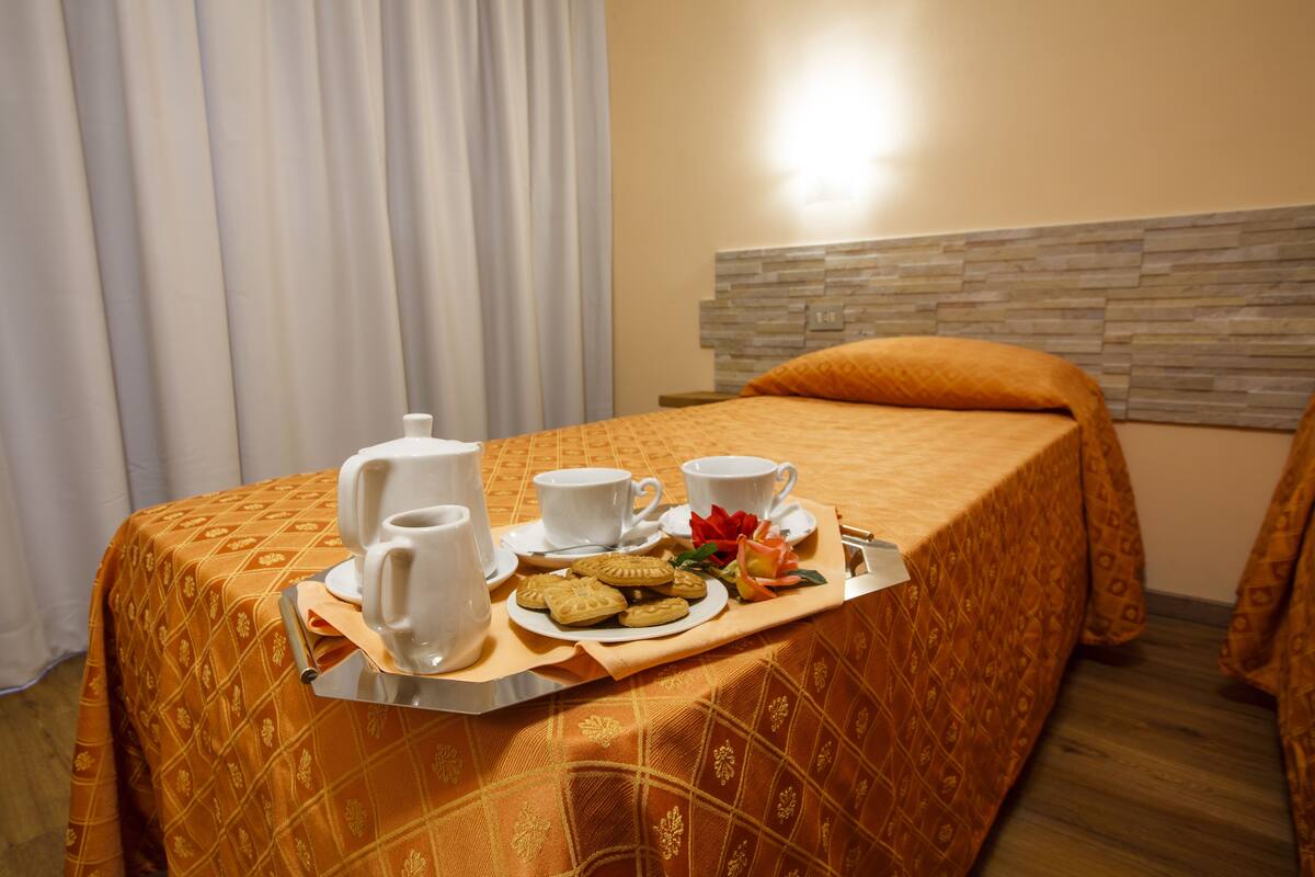 Un lit bien fait, recouvert d’une couvre-lit à motifs, un plateau avec des éléments de petit-déjeuner, notamment des biscuits, une théière, des tasses et une rose.