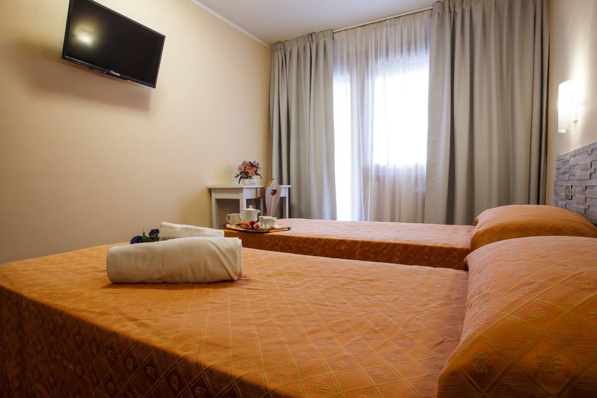 Une chambre d’hôtel avec deux lits, un téléviseur à écran plat fixé au mur, une petite table avec un vase de fleurs et une fenêtre avec des rideaux.