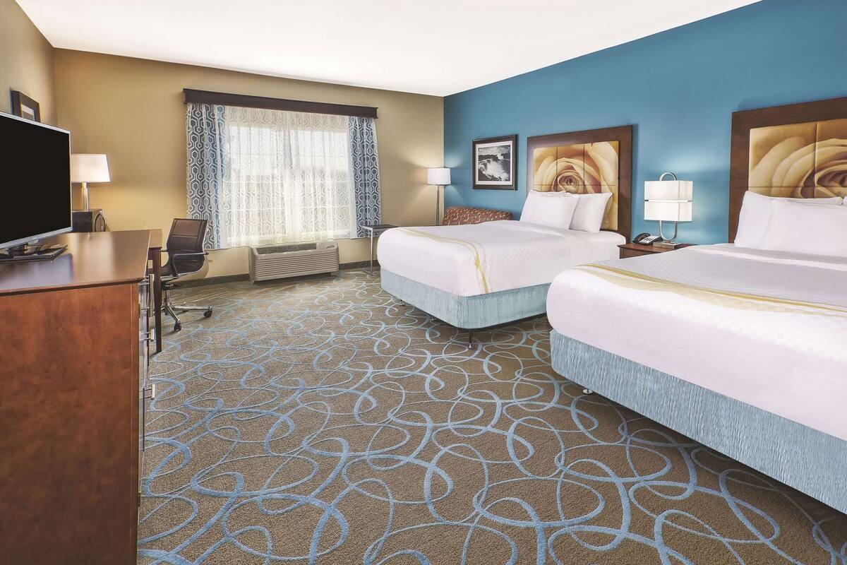 Ein Hotelzimmer mit zwei Betten, einem Schreibtisch, einem Fernseher und einem Fenster mit gemusterten Vorhängen.
