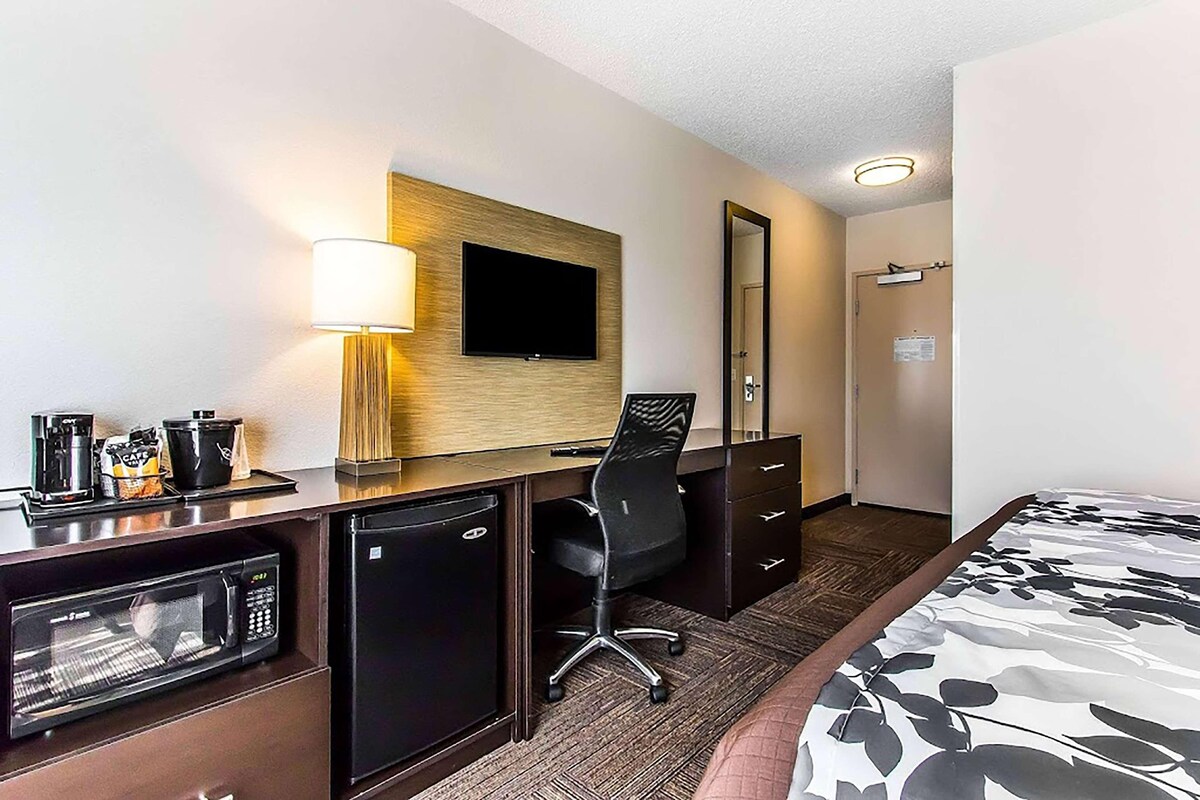 A hotel room with a bed, desk, microwave, refrigerator, chair, and a TV mounted on the wall.